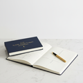 Nocturely Sleep Tracking Journal deep navy hardcover with gold foil lettering open on white marble surface with a pen